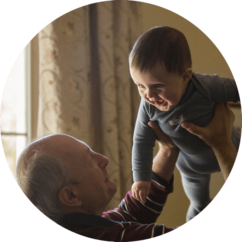 A grandfather is shown smiling as he lifts his grandchild up in the air while the baby giggles.