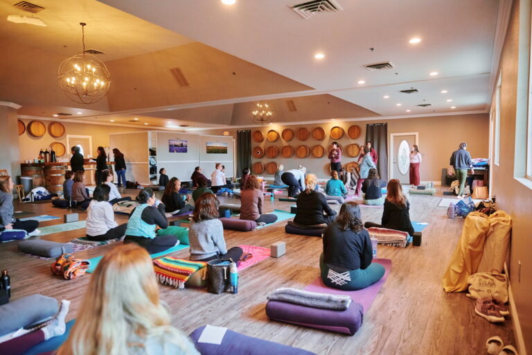A group of women sitting on yoga mats in a large room with teachers standing at the front